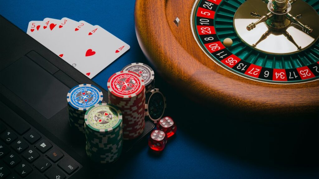 Close-up of poker chips, playing cards, and a roulette wheel next to a laptop in a casino setting